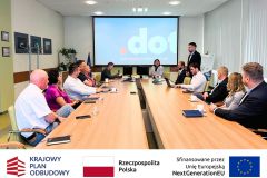 Spotkanie, przy stole siedzą uczestnicy po lewej i prawej stronie stołu, w tle ekran z prezentacją, jeden mężczyzna stoi i przemawia.