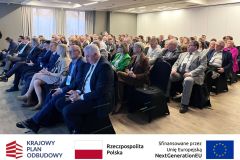 Uczestnicy konferencji. Po zdjęciem belka z logotypami KPO, flaga Polski oraz flaga Unii Europejskiej i napisem "Sfinansowano przez Unie Europejską".