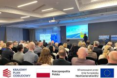 Sala konferencyjna z uczestnikami, do których przemawia prelegent. W tle prezentacja oraz rollupy. Po zdjęciem belka z logotypami KPO, flaga Polski oraz flaga Unii Europejskiej i napisem "Sfinansowano przez Unie Europejską".