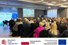 Sala konferencyjna, uczestnicy przysłuchują się prezentacji. Po zdjęciem belka z logotypami KPO, flaga Polski oraz flaga Unii Europejskiej i napisem "Sfinansowano przez Unie Europejską".