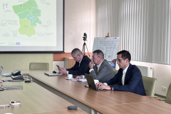 Trzech mężczyzn siedzi przy stole konferencyjnym, w tle prezentacja multimedialna.