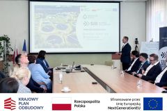 Sala konferencyjna, przy stole siedzą uczestnicy, 10 osób, jedna prezentuje. W tle prezentacja multimedialna. Po zdjęciem belka z logotypami KPO, flaga Polski oraz flaga Unii Europejskiej i napisem "Sfinansowano przez Unie Europejską".