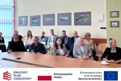 Sala konferencyjna, uczestnicy siedzą w dwóch rzędach: jedni przy stole, drudzy za nimi, przysłuchują się. Po zdjęciem belka z logotypami KPO, flaga Polski oraz flaga Unii Europejskiej i napisem "Sfinansowano przez Unie Europejską".