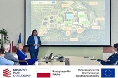 Sala konferencyjna, przy stole siedzą uczestnicy, 10 osób, jedna prezentuje. W tle prezentacja multimedialna. Po zdjęciem belka z logotypami KPO, flaga Polski oraz flaga Unii Europejskiej i napisem "Sfinansowano przez Unie Europejską".