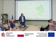 Sala konferencyjna, przy stole siedzą uczestnicy, kobiety i mężczyźni. W tle prezentacja multimedialna.Po zdjęciem belka z logotypami KPO, flaga Polski oraz flaga Unii Europejskiej i napisem "Sfinansowano przez Unie Europejską".