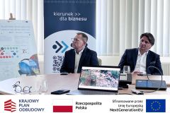 Dwóch mężczyzn siedzi przy stole, przysłuchują się prezentacji, za nim rollup Słupskiej Izby Biznesu oraz tablica informacyjna projektu SWB. Na stole laptop, mikrofon, okulary. Po zdjęciem belka z logotypami KPO, flaga Polski oraz flaga Unii Europejskiej i napisem "Sfinansowano przez Unie Europejską".