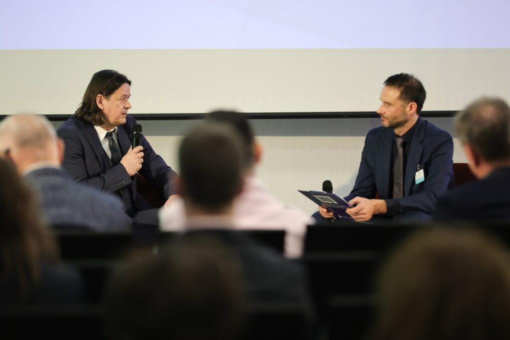 Dwóch mężczyzn siedzi i rozmawia w ręku trzymają mikrofony, obserwują ich uczestnicy konferencji.